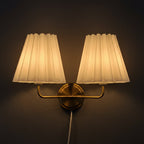 Double-head wall sconce with two pleated cream shades, gold bracket, visible cord